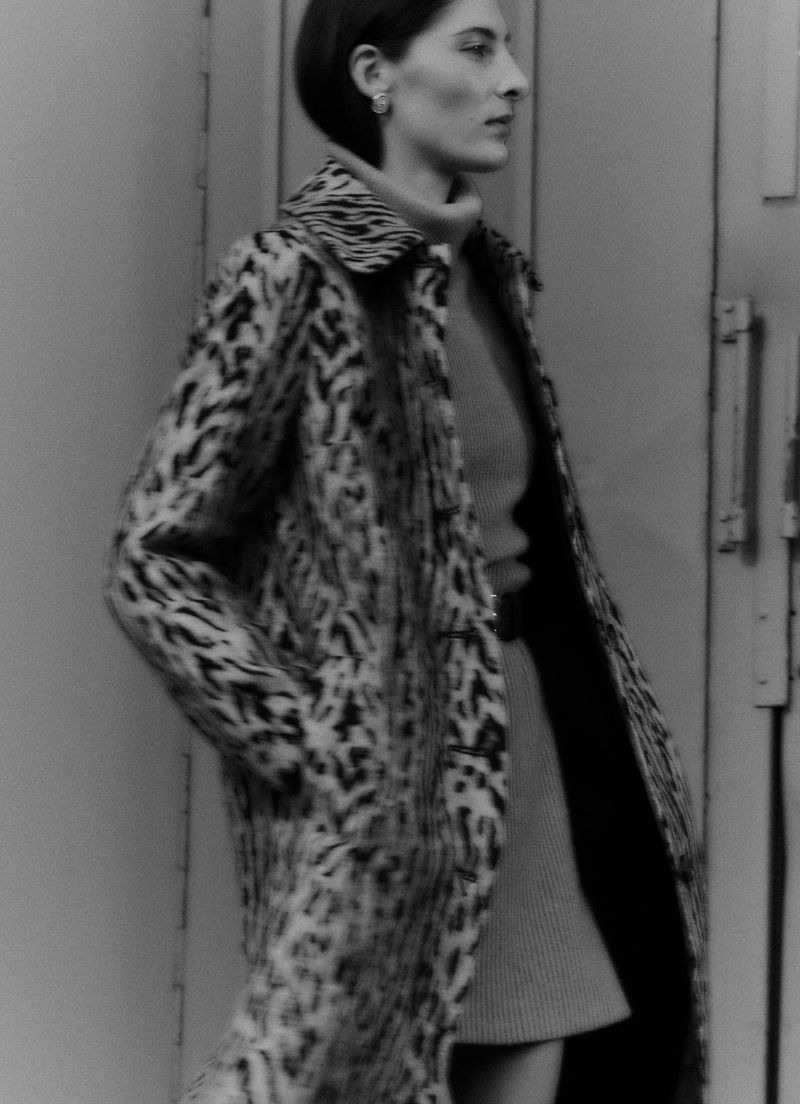 A woman in a patterned coat poses near a gray wall. The image is a monochrome fashion shot with a shallow depth of field.