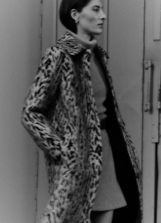 A woman in a patterned coat poses near a gray wall. The image is a monochrome fashion shot with a shallow depth of field.