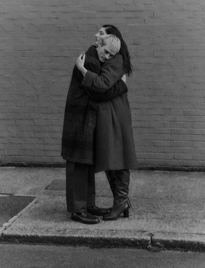 Two people embracing in front of a brick wall; a person with a long dark coat is hugging another person with a long light coat. 