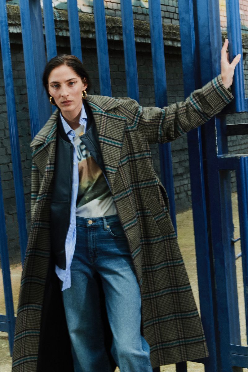 Woman in a plaid coat standing next to a blue metal gate, wearing jeans and a graphic tee in an urban environment. Fashion shot.
