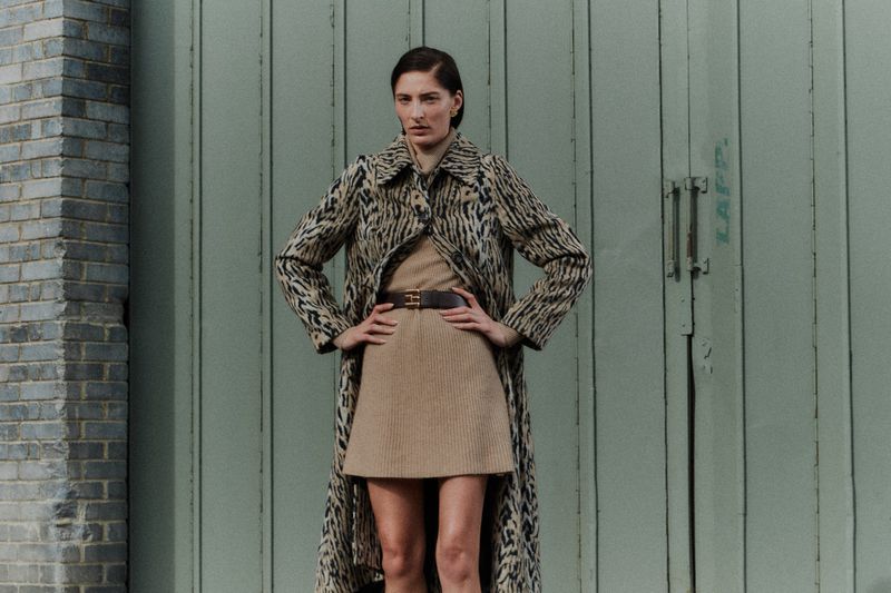 A woman poses in a patterned coat and neutral dress, standing confidently against a weathered wall and blue-green door backdrop.