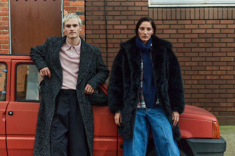 Two people in winter coats pose next to an orange car against a brick wall, showcasing fashion and automotive style in an outdoor setting.