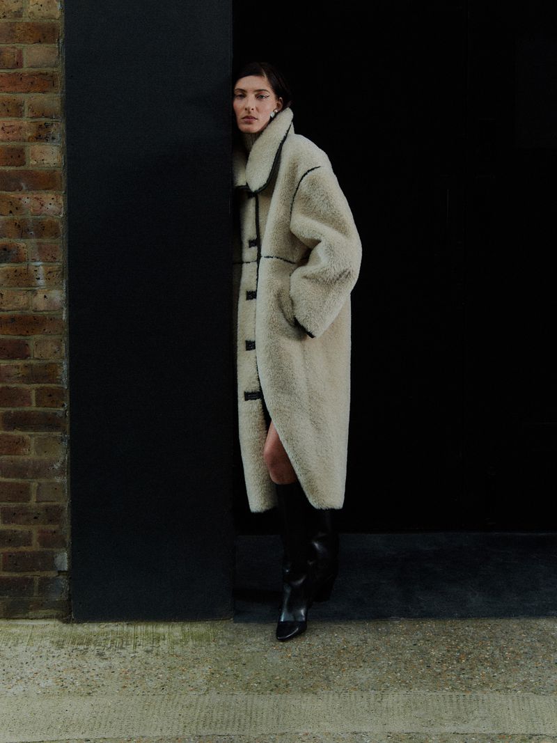 A woman poses in a long cream-colored sherpa coat and black boots in a gritty urban setting with dark walls and concrete flooring.