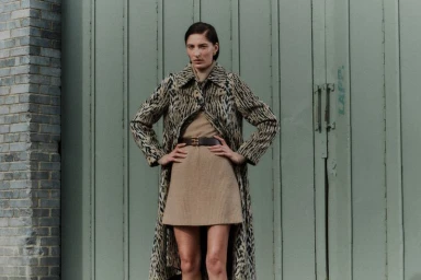 Model wearing a patterned animal print coat over a beige dress against green wooden doors for The Observer Magazine UK.