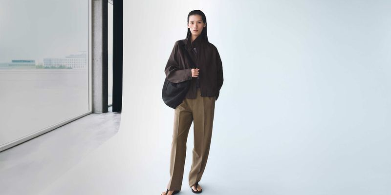 Full-length shot of a woman in brown jacket and trousers posing in a studio with a window, minimalist fashion photography.