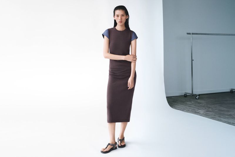 An adult woman stands in a photo studio, wearing a brown dress with blue cap sleeves and dark platform sandals, against a white background.