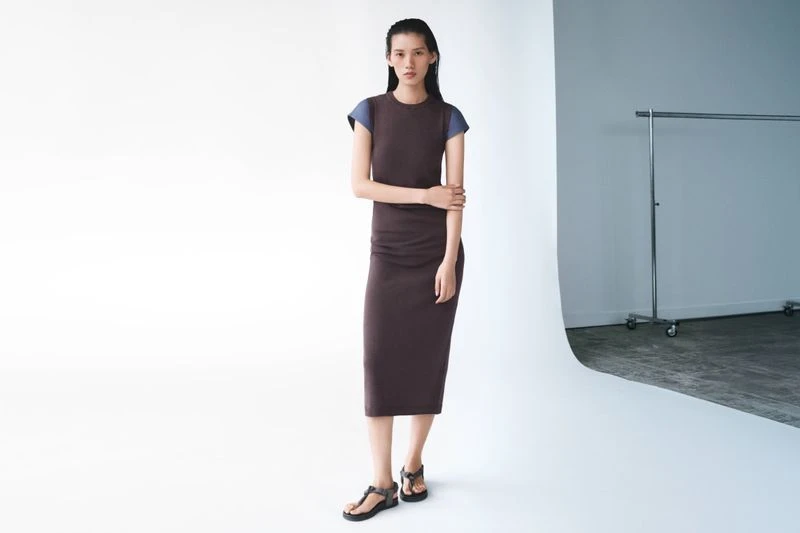 An adult woman stands in a photo studio, wearing a brown dress with blue cap sleeves and dark platform sandals, against a white background.