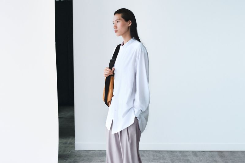 Fashionable Asian woman in white shirt and trousers carrying a tan bag, posing in a white studio with soft natural light, looking away.