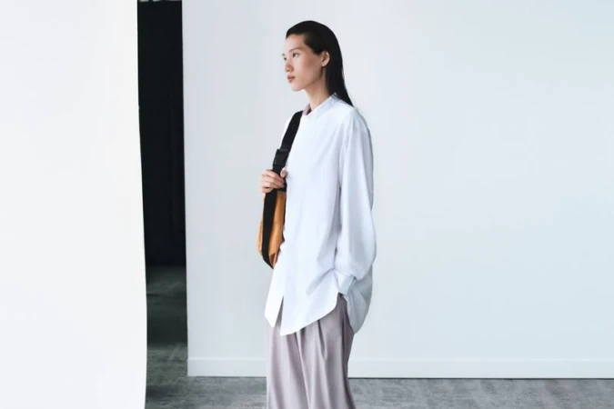 Fashionable Asian woman in white shirt and trousers carrying a tan bag, posing in a white studio with soft natural light, looking away.