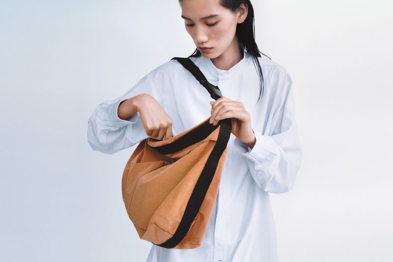 A woman in a white shirt looks into a brown hobo bag with a black strap in a studio shot against a white backdrop.