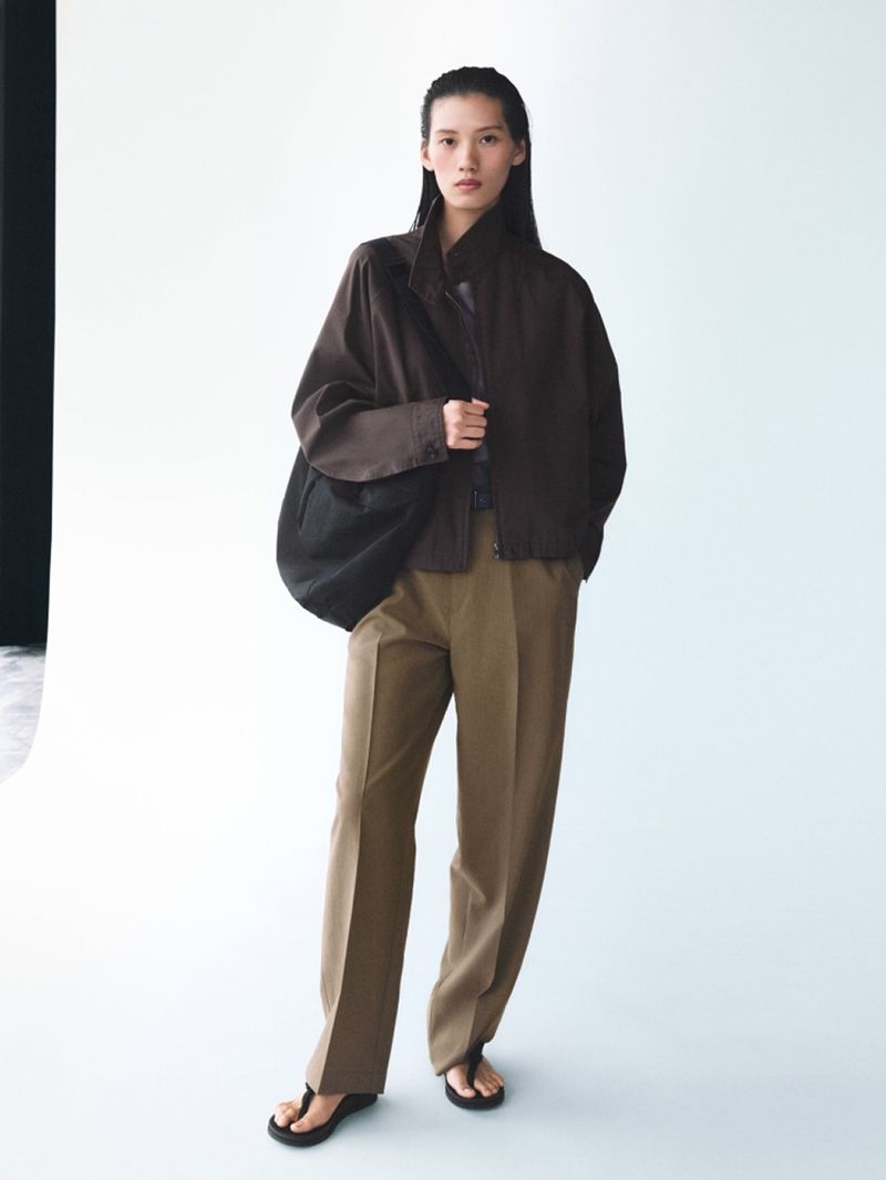 Full-length shot of an Asian fashion model posing indoors, wearing a brown jacket, khaki trousers, black shoulder bag, and sandals.