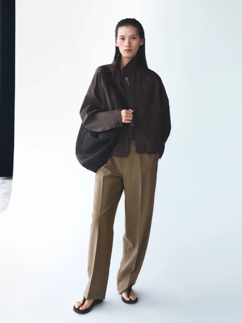 Full-length shot of an Asian fashion model posing indoors, wearing a brown jacket, khaki trousers, black shoulder bag, and sandals.