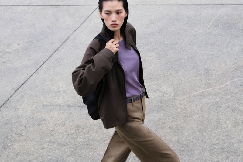 A slender woman with a backpack walks on concrete wearing a purple top, jacket, and brown pants in an outdoor fashion shot.