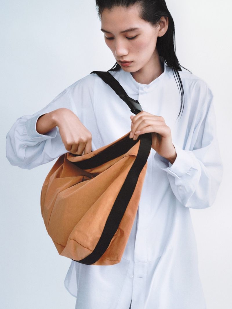 Studio shot of a woman modeling a brown hobo bag with a black strap, posing against a white background in a long-sleeved blue shirt.