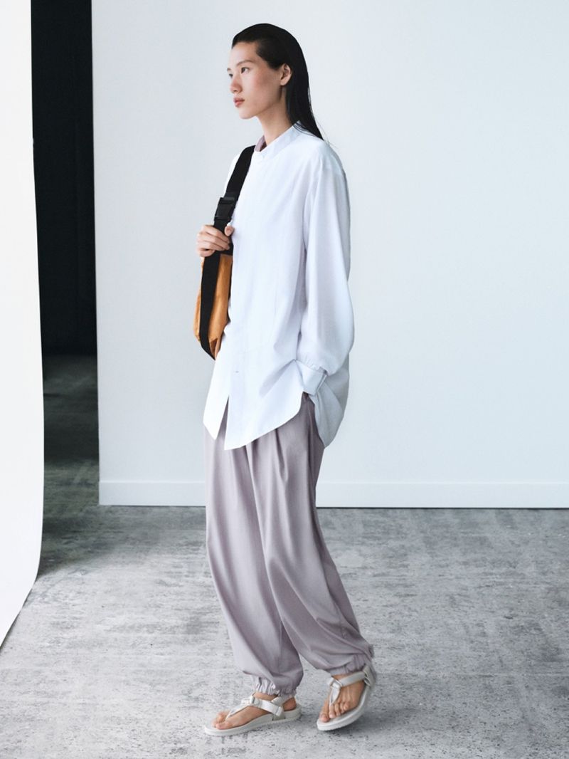 Side profile shot of an Asian woman in a white shirt and grey pants with a brown bag, in a studio with a white wall.