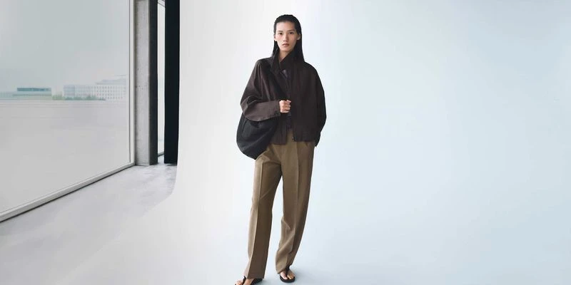 Model Jiayun Xie wearing a dark brown jacket and trousers carrying a black shoulder bag in front of a white studio sweep backdrop.
