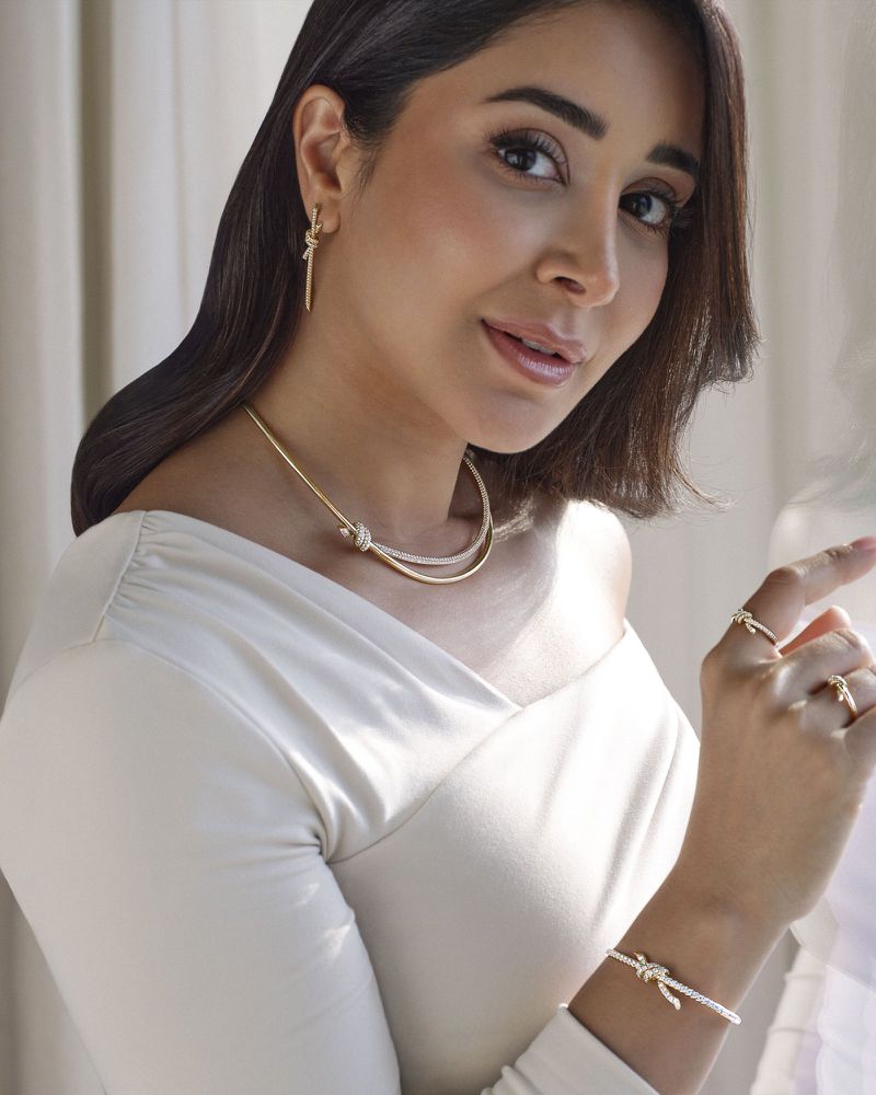 Woman wearing a white long-sleeve shirt, jewelry, and gold choker poses in a close-up, portrait shot with soft studio lighting.