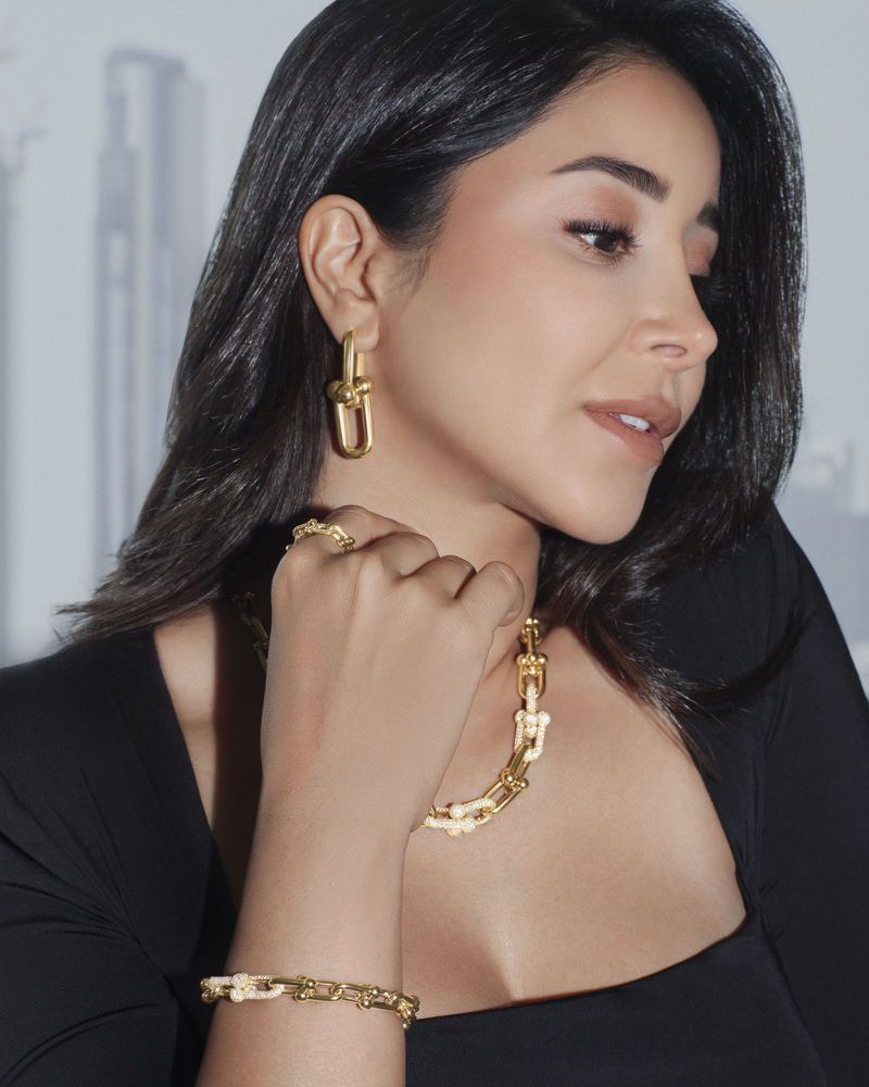 Close-up studio shot of a woman wearing a black top and gold jewelry, including statement earrings, a necklace, a ring, and a bracelet.
