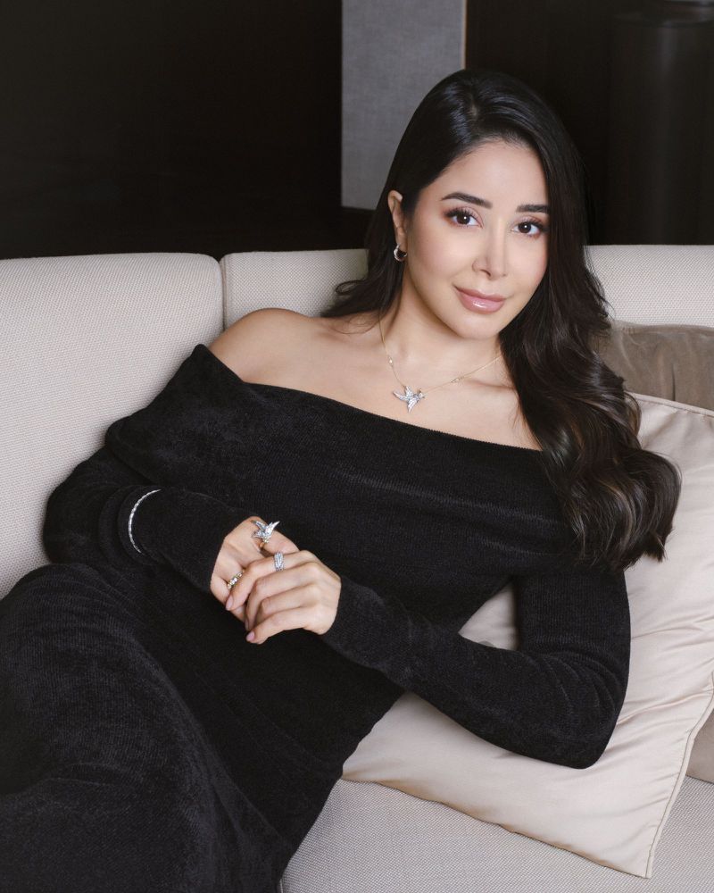 Elegant woman in black off-shoulder dress with jewelry, sitting on a beige sofa, in an indoor setting, posing for a portrait.