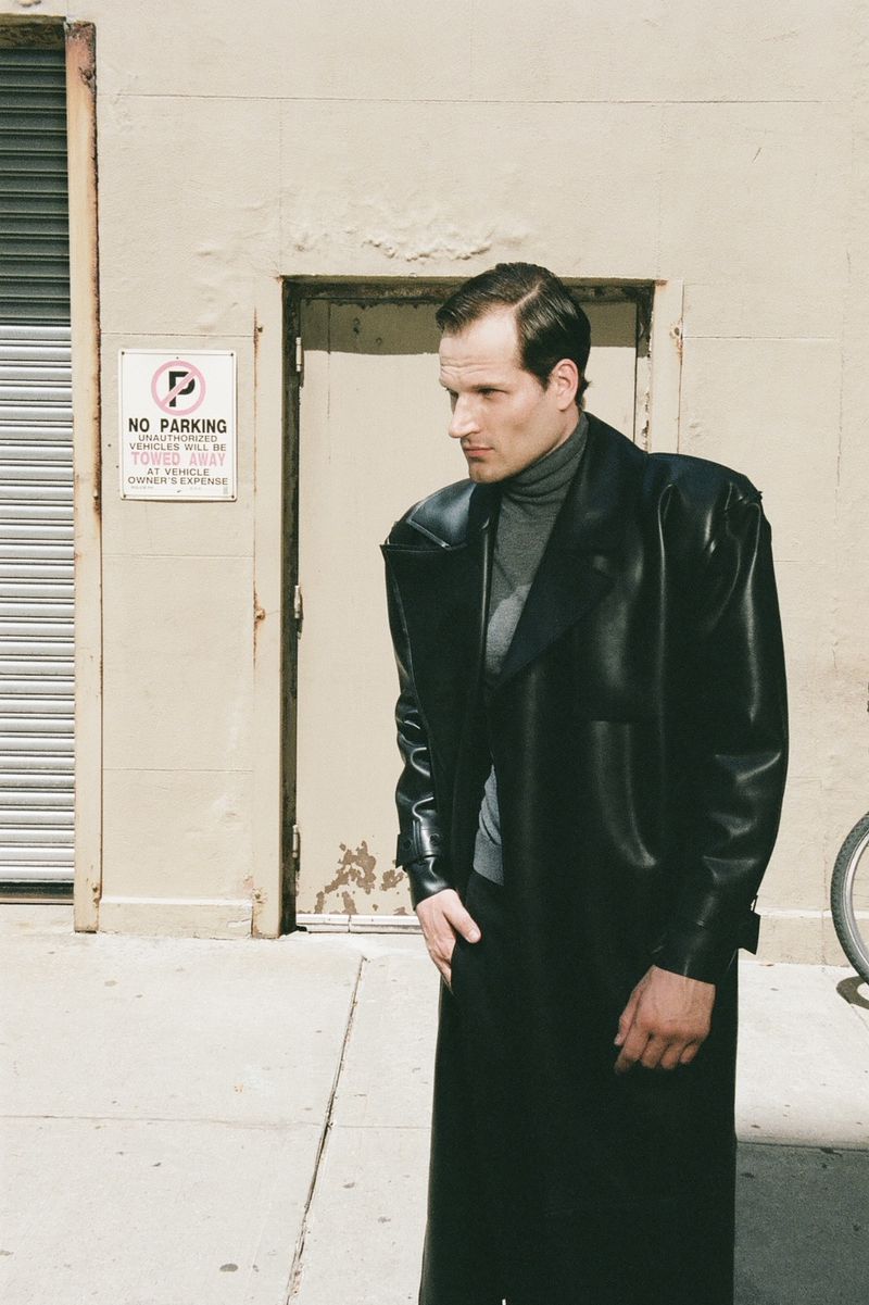 Adult male in a long black leather coat standing on a city street. Neutral facial expression, gray turtleneck, and an urban setting.
