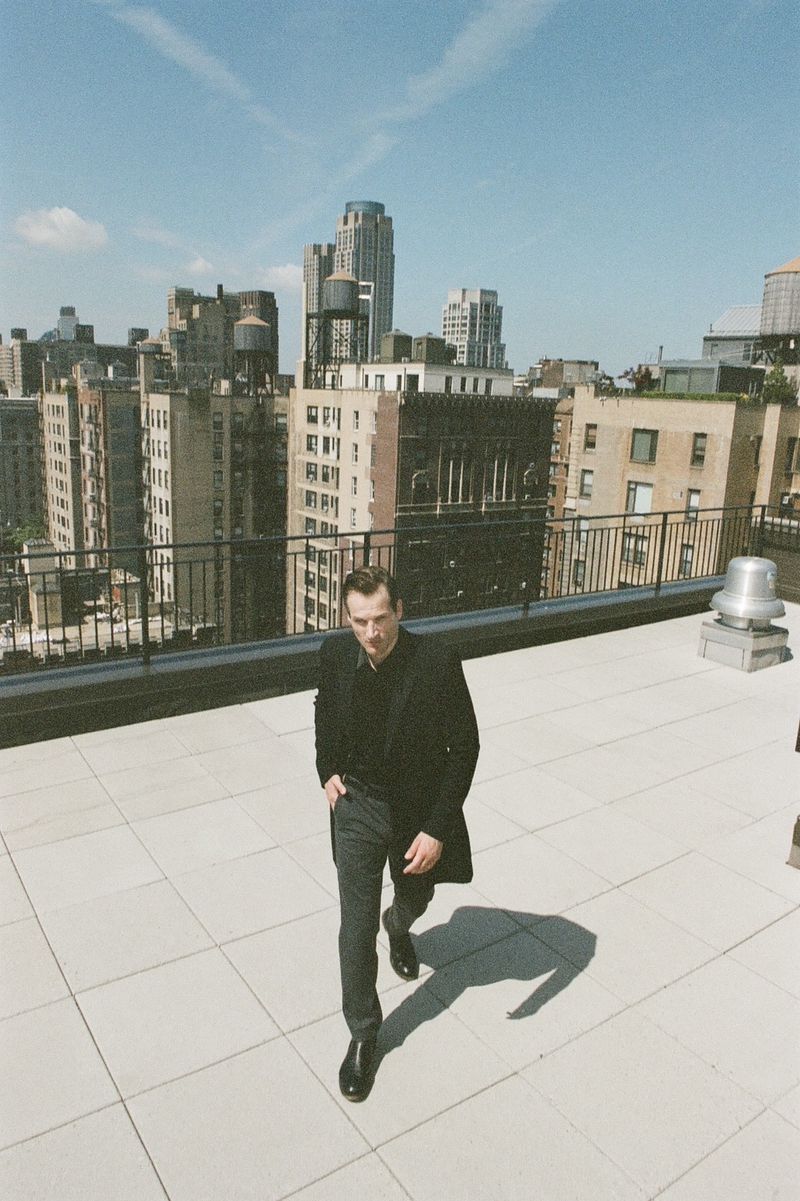 Man in a black coat walks on a rooftop with city views. Posing against a backdrop of buildings under a clear sky.