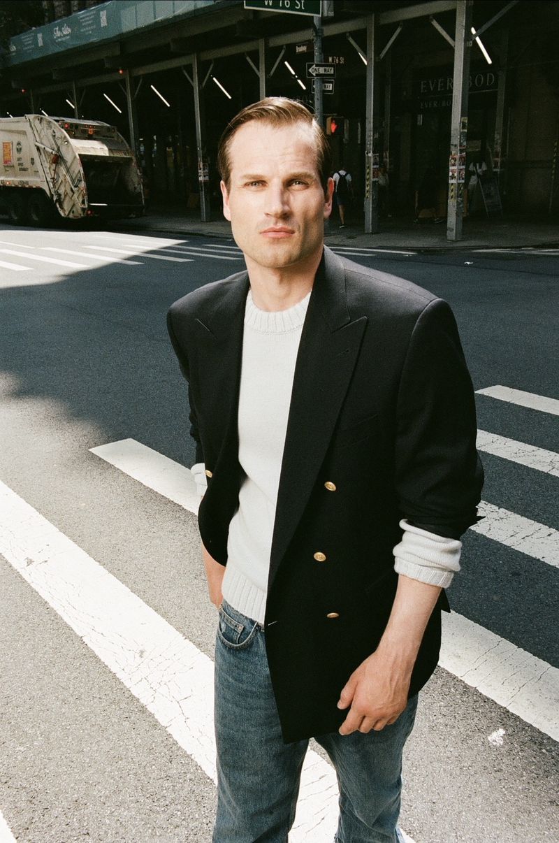 Adult male in black blazer and jeans stands on a crosswalk in an urban setting, with a garbage truck and buildings visible.