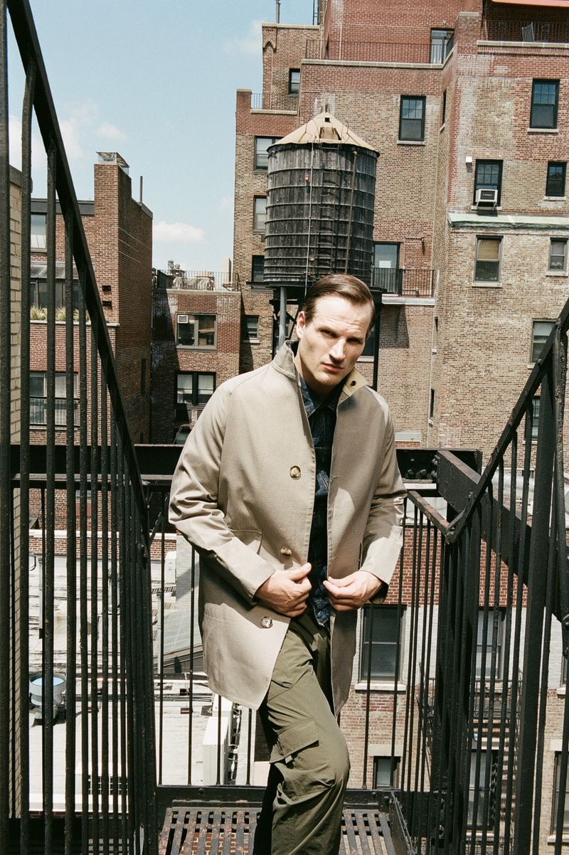 Man stands on metal stairs on a city rooftop, wearing a trench coat, button shirt, and cargo pants, with brick buildings around him.