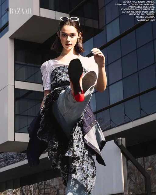 Fashion editorial shot of a young adult woman in a patchwork denim dress, posing outdoors against modern architecture with Bazaar logo.