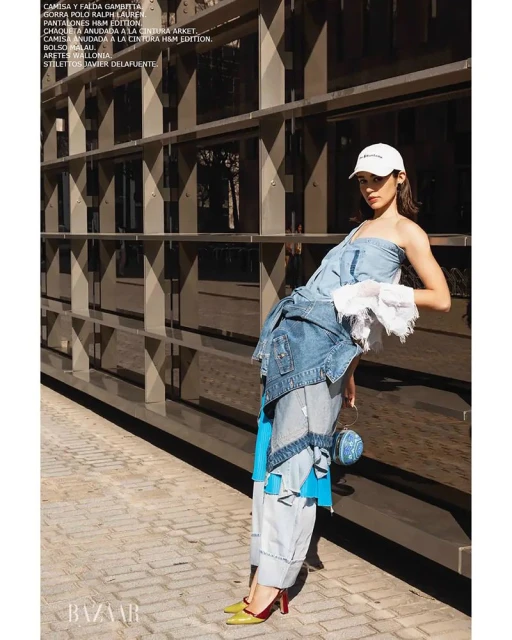 Fashion editorial shot of a woman wearing a patchwork denim outfit with a cap and heels, leaning against a modern metal wall.