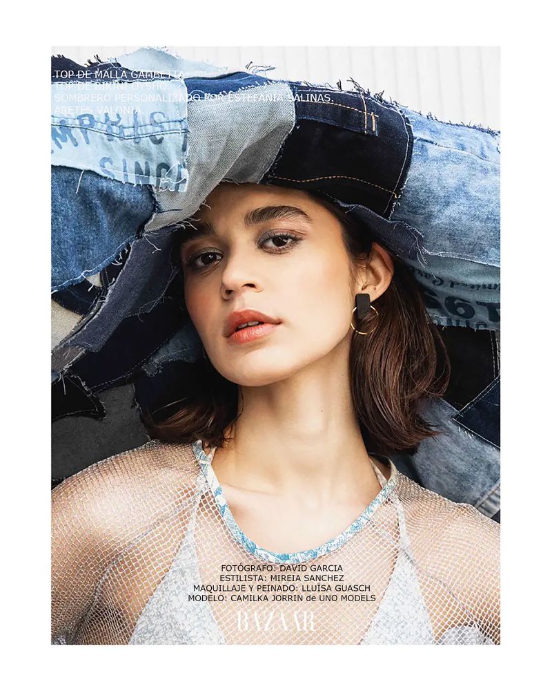Fashion shot of a female model wearing a denim patchwork hat and mesh top, posing against a light backdrop with natural lighting.