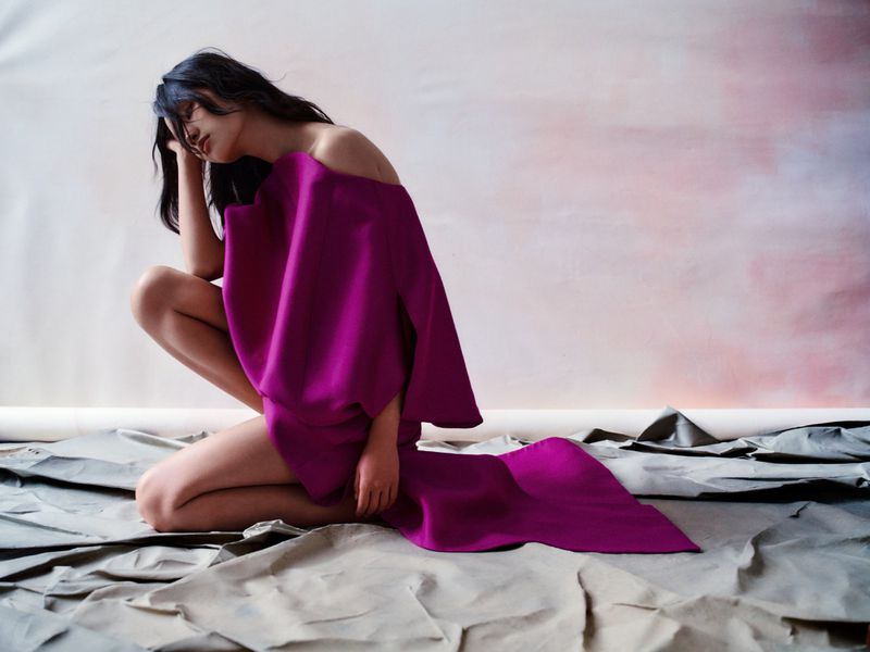Woman in a magenta dress crouches indoors on a tan cloth, posing against a pink-tinted wall in a studio environment with soft lighting.