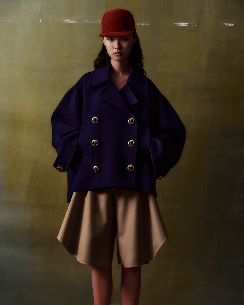 A stylish Asian woman models a navy double-breasted jacket with gold buttons and beige shorts in a fashion studio setting.
