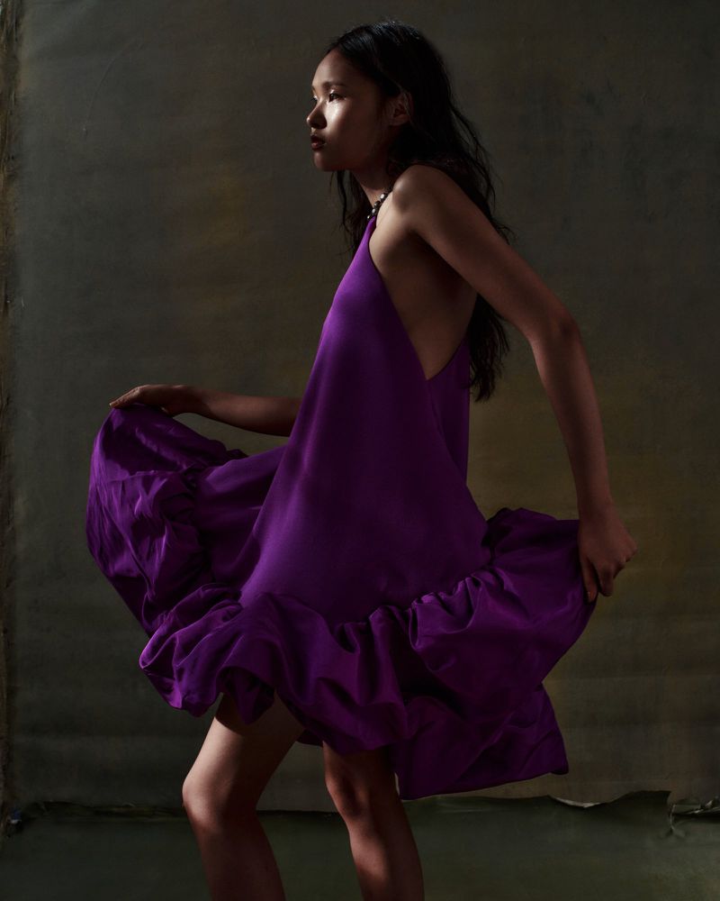 Fashion shot of an Asian woman posing in a purple dress against a textured neutral background, side view, medium shot, studio lighting.