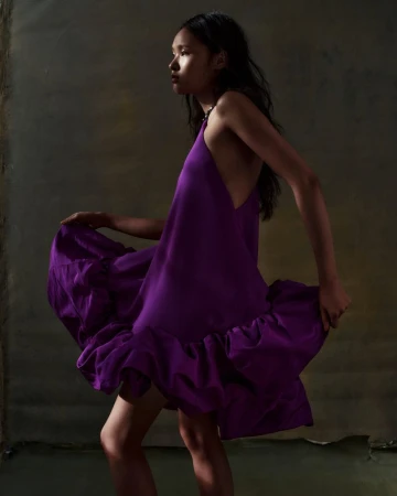 Fashion shot of an Asian woman posing in a purple dress against a textured neutral background, side view, medium shot, studio lighting.