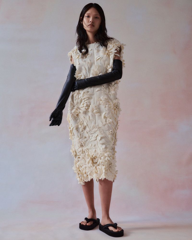 An Asian fashion model poses in a sleeveless, flower-decorated dress with long black gloves and platform sandals in a studio setting.