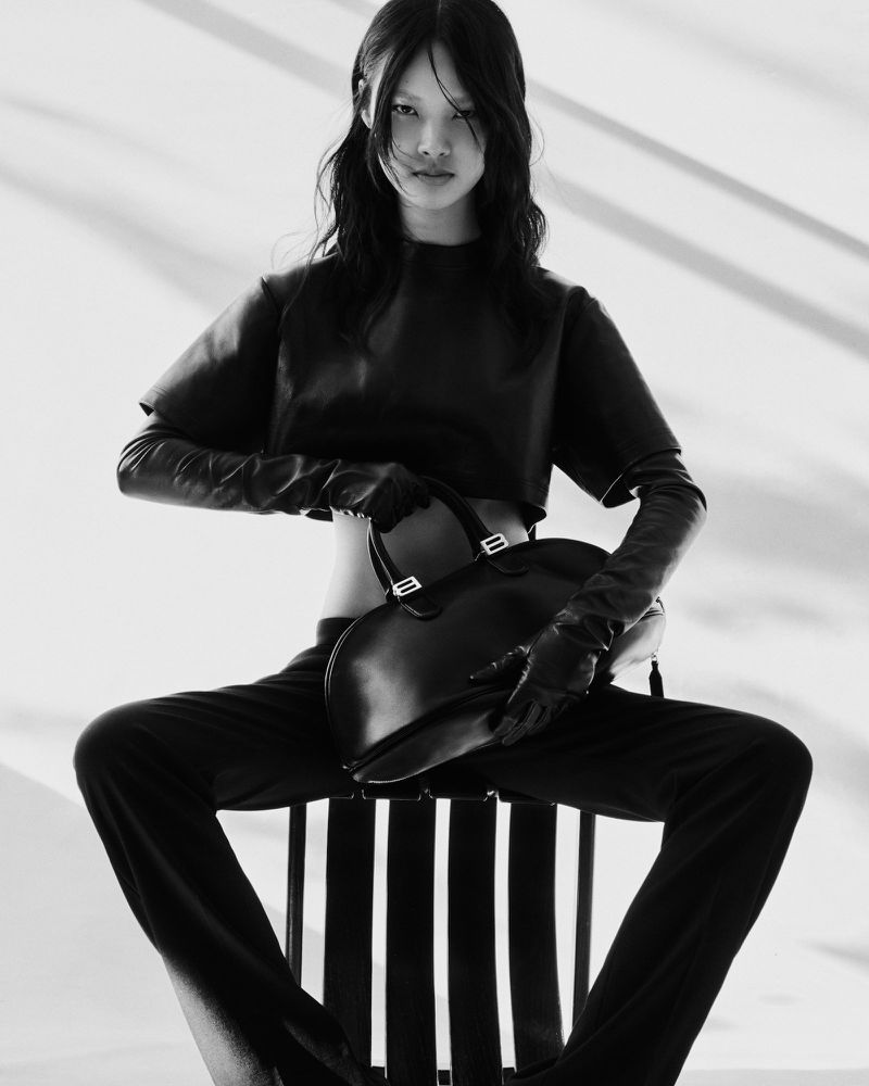 Black-and-white portrait of an Asian woman with long hair wearing a black crop top and pants, sitting on a chair while holding a black bag.