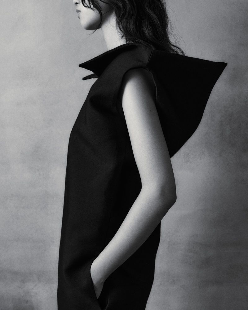 Monochrome fashion shot: adult female model poses in a black sleeveless dress with a large collar against a neutral background in a photo studio.