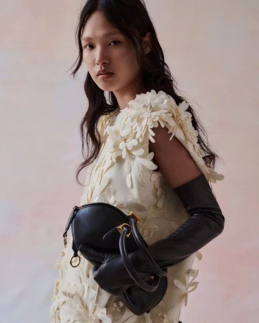 Asian woman in cream floral dress holds black purse wearing long black gloves in a studio, fashion editorial for Visie.