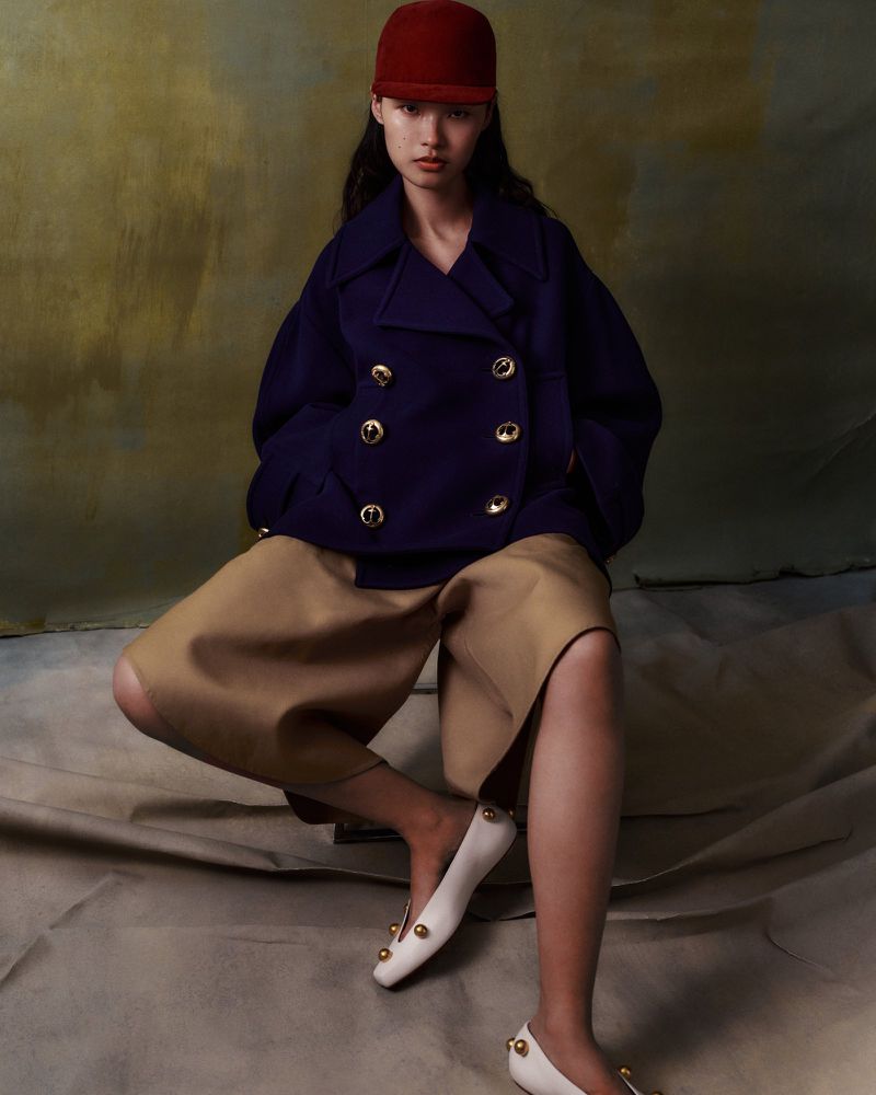 Model wearing a navy peacoat, tan shorts, red hat, and white flats sitting against a textured backdrop