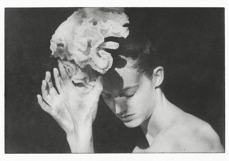 Black and white portrait of a woman with a flower on her head, eyes closed, hands near face against a dark background.