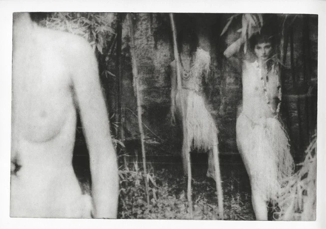 Monochrome photograph featuring a woman in a decorative outfit, standing in a jungle-like setting with foliage and branches.