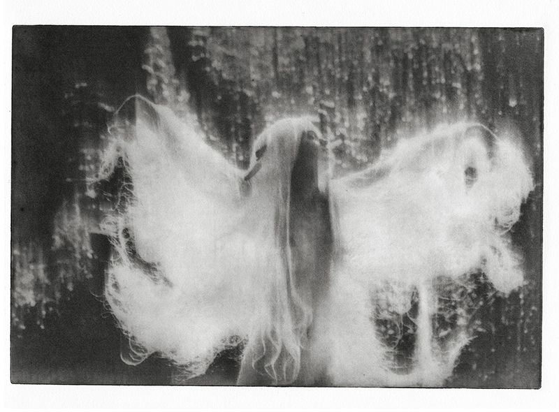 Monochromatic fine art photo of blurred figure in flowing cloth before dappled background, creating ethereal and abstract image.