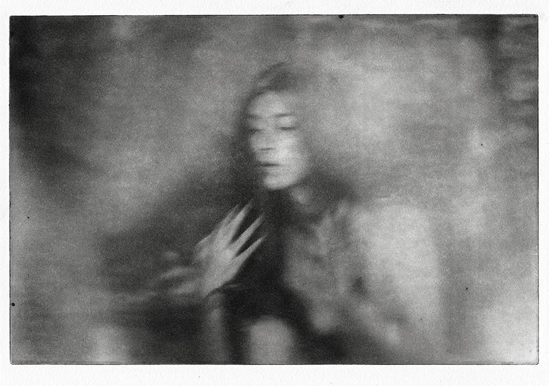 Monochrome portrait of a woman with a serene expression, her hand near her chest, captured in a soft-focus, dreamlike artistic style.