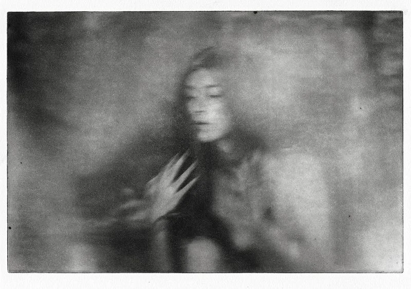 Monochrome portrait of a woman with a serene expression, her hand near her chest, captured in a soft-focus, dreamlike artistic style.