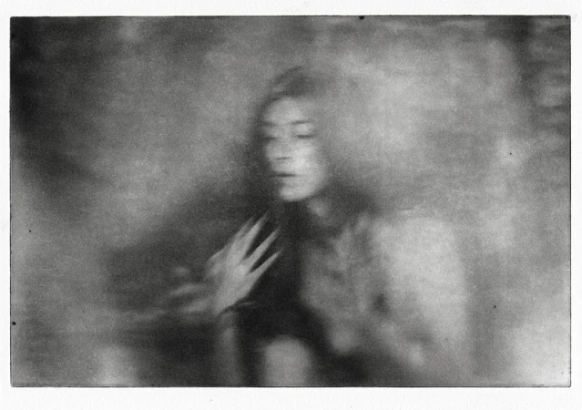 Monochrome portrait of a woman with a serene expression, her hand near her chest, captured in a soft-focus, dreamlike artistic style.