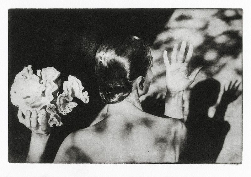 Monochrome art photography: Woman with hand up, holding coral, casting shadow on a textured backdrop with back lighting.