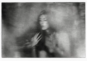 Monochrome motion-blurred portrait of a woman with a hand raised near her chest against a textured, grainy background.