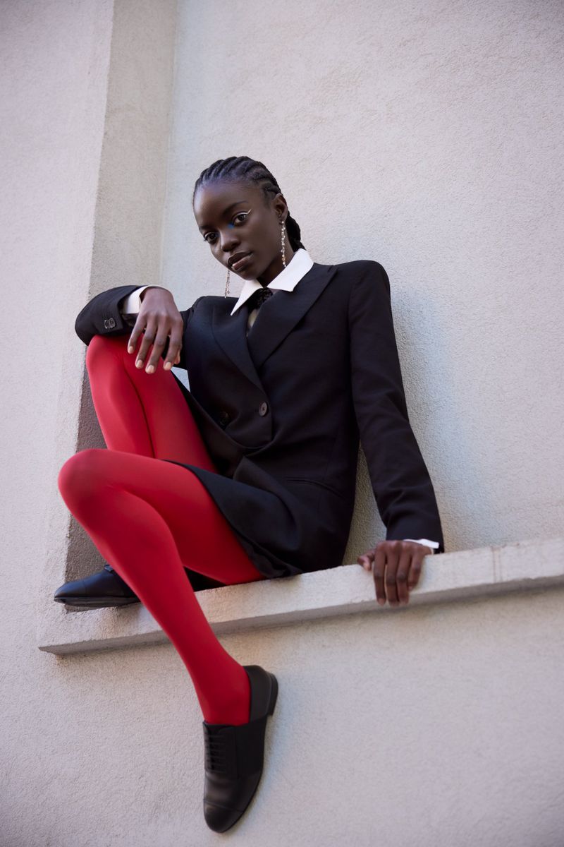 Portrait of a dark-skinned woman in a black suit with red tights sitting outdoors with a serious expression, looking at the camera.