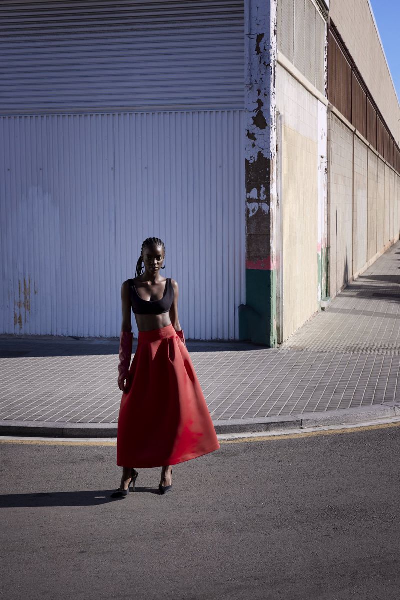 Woman in black top and red skirt poses on urban street corner near a building, captured in natural light with a medium shot.