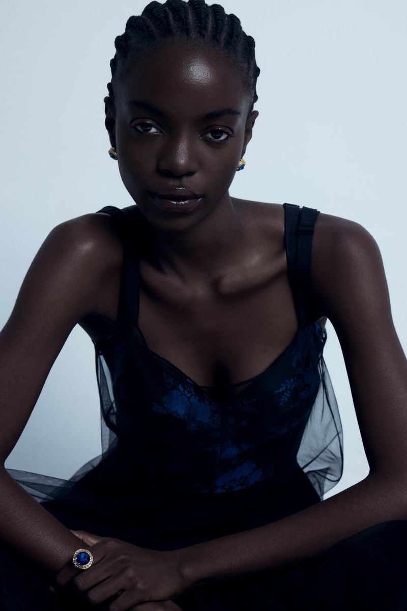 Portrait of a Black woman with braided hair, wearing a black top and jewelry, posing in a studio against a white backdrop.